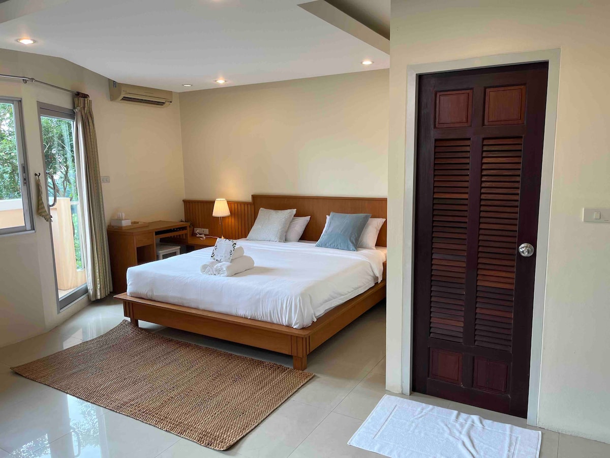 A spacious bedroom features a large bed with neutral linens and two pillows. A small wooden desk and lamp are positioned nearby, while large windows allow natural light to fill the room. A woven rug adds warmth to the tiled floor.
