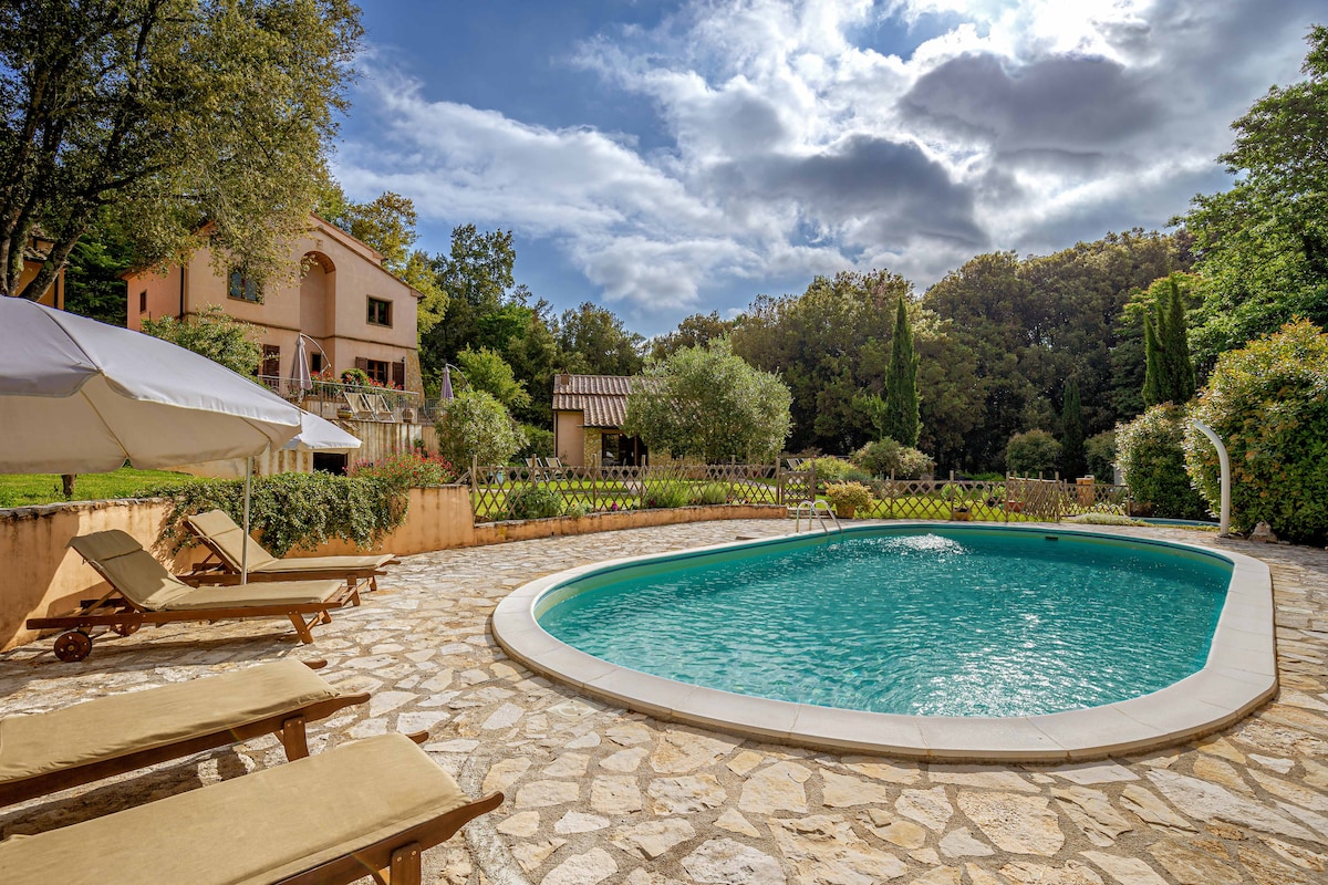 A swimming pool is surrounded by stone decking, with several pool loungers positioned nearby. Lush greenery and trees are visible in the background, while a house with a terracotta façade is nestled among the foliage, providing a serene outdoor space.