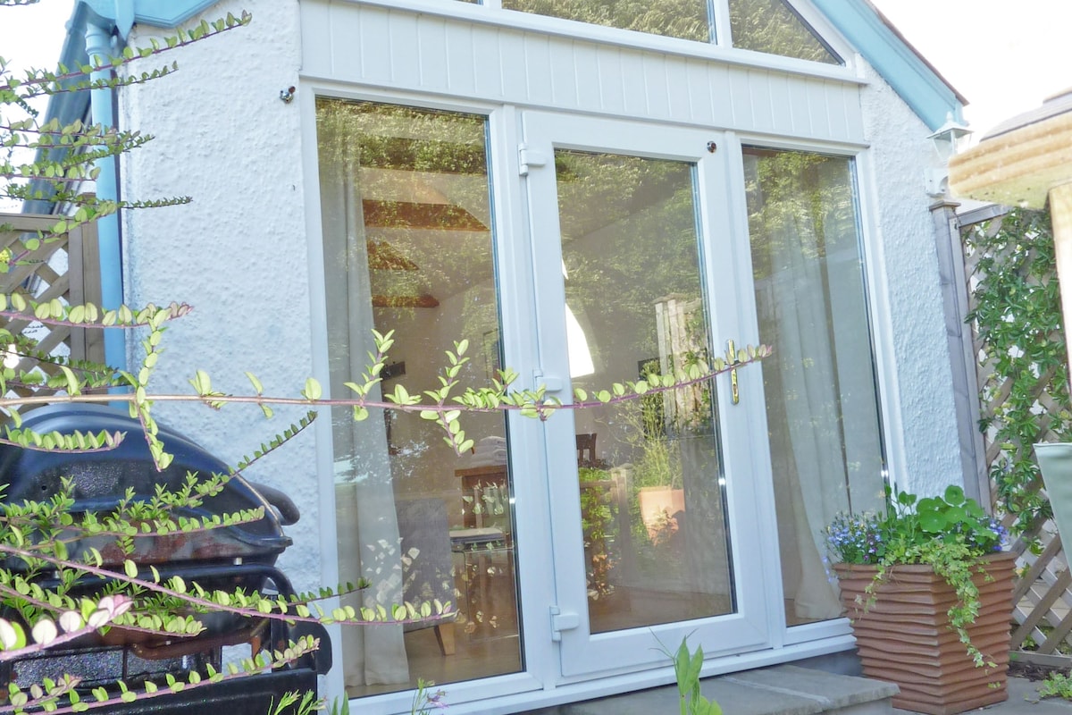 The exterior of the Boathouse is viewed, showcasing large glass doors that connect the indoor and outdoor spaces. Lush greenery surrounds the entrance, and a barbecue grill is positioned nearby, inviting outdoor dining options.