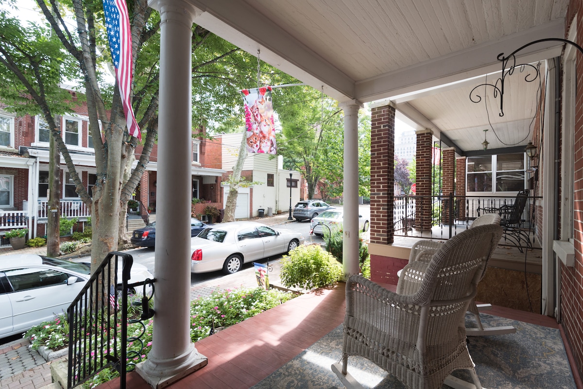 A charming porch area is revealed, featuring wicker rocking chairs and decorative elements. Lush greenery surrounds the entrance, and an inviting view of the tree-lined street is visible, showcasing parked vehicles and neighboring homes. Soft morning light filters through the trees, enhancing the serene setting.