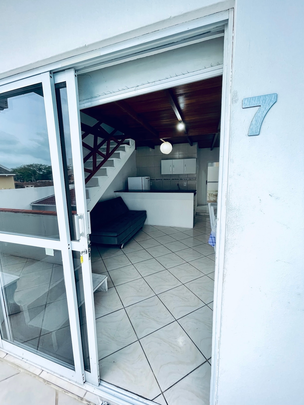 A view from the entrance showcases an open-plan living space with a sofa bed and a kitchen area. Natural light enters through glass doors, illuminating the tiled floor. A staircase leads to the upper level, where additional sleeping arrangements are located.