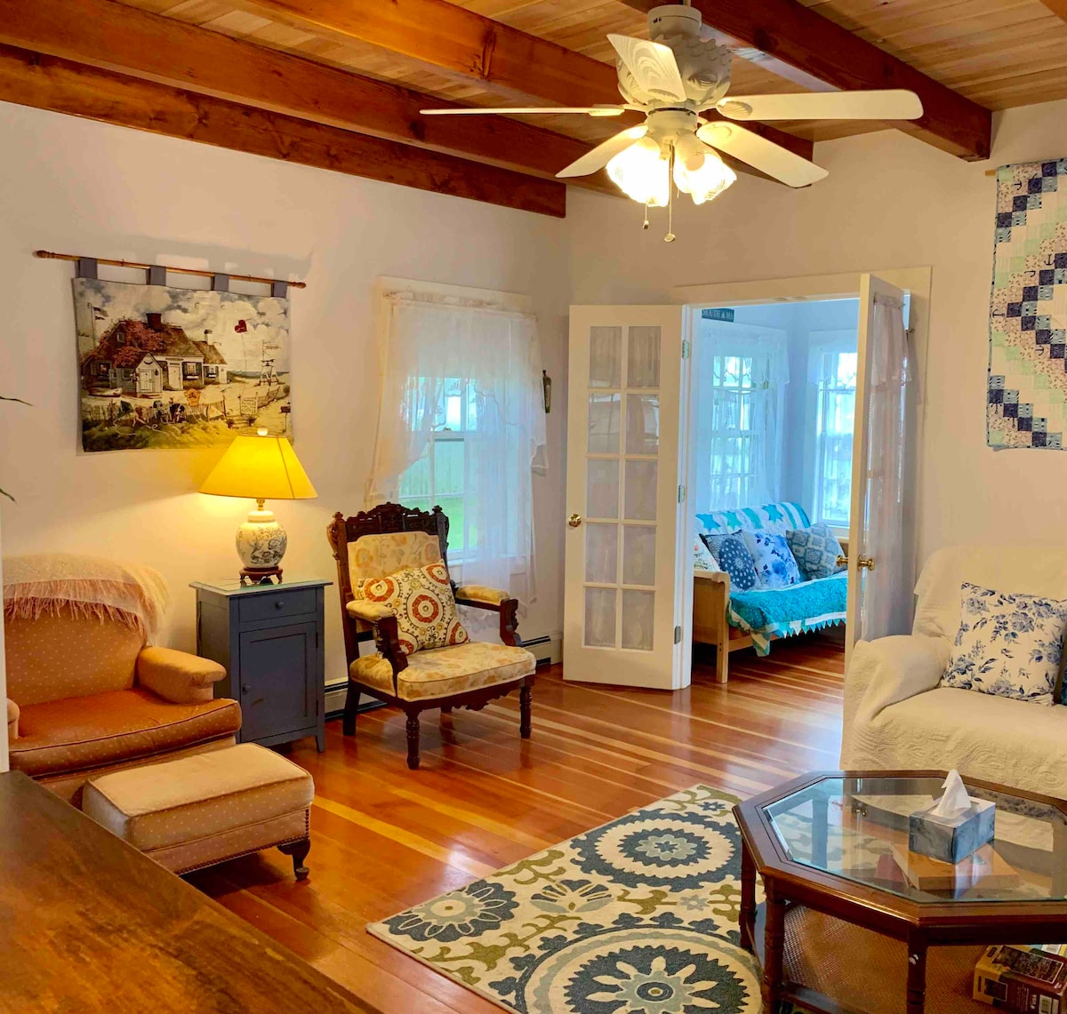 A welcoming living area is highlighted by wooden beams and natural light. Comfortable seating options include a pastel armchair and patterned upholstered chair. A glass-topped coffee table rests on a decorative area rug. The room features a light fixture and a painting on the wall.
