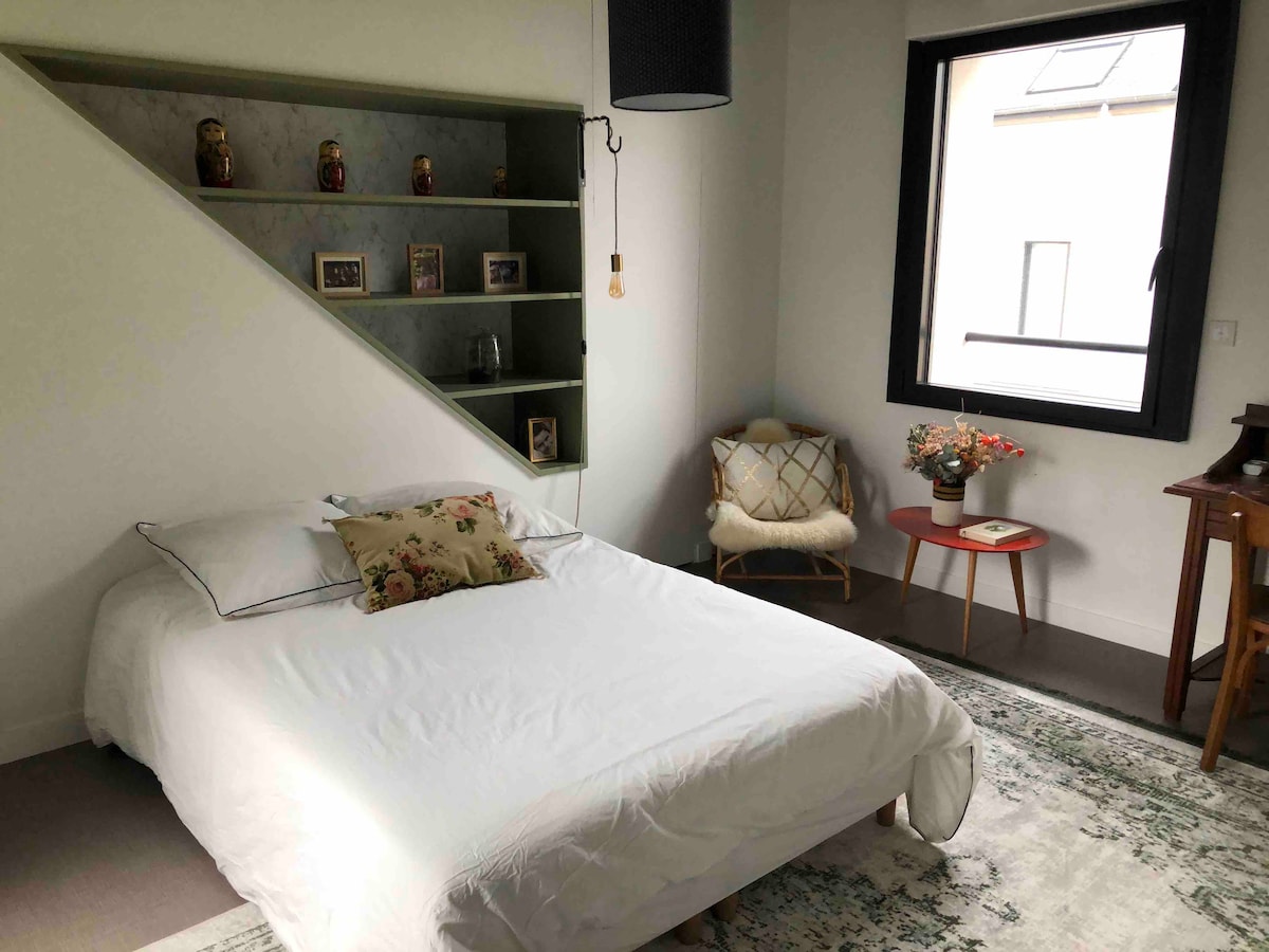 A bright and airy bedroom features a spacious bed adorned with decorative pillows. A set of shelves with various items is positioned nearby, and a comfortable chair sits beside a small side table. Natural light enters through the large window, enhancing the room's tranquility.
