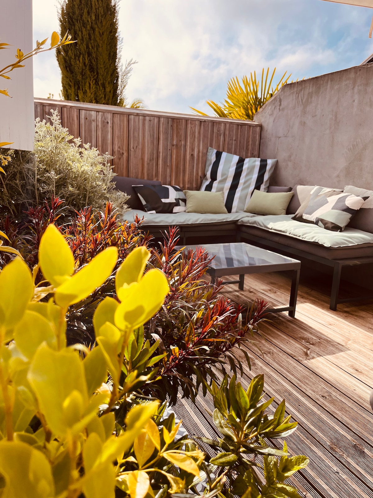 A comfortable outdoor lounge area is depicted, adorned with various decorative pillows. A low table sits on a wooden deck, surrounded by vibrant green and yellow foliage, creating a relaxing atmosphere. The backdrop includes a wooden fence and hints of blue sky.
