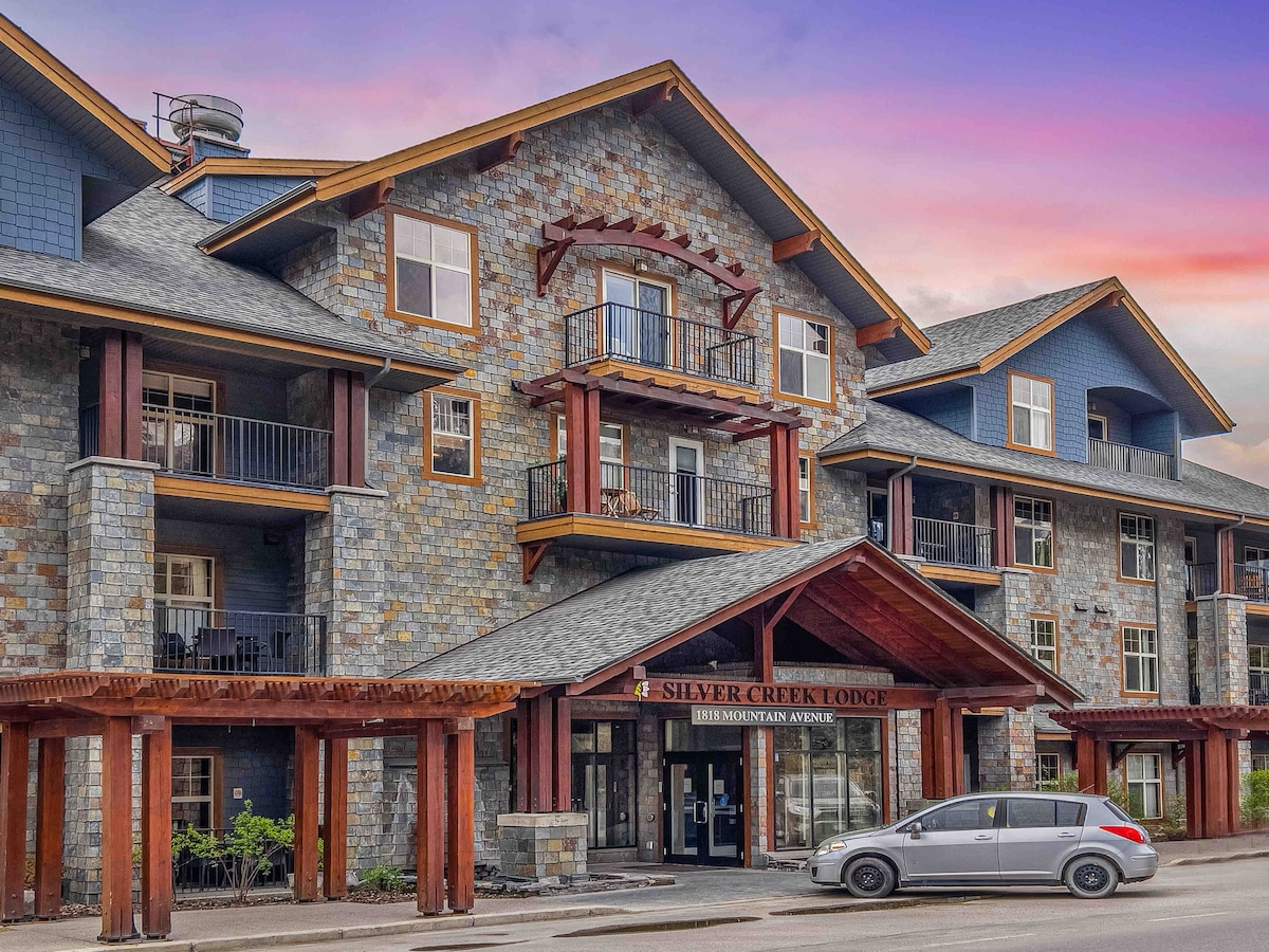 The exterior of Silver Creek Lodge is showcased, featuring stone and wood accents. The three-story building includes balconies that overlook the entrance. A single vehicle is parked in front, and a colorful sky is visible in the background.