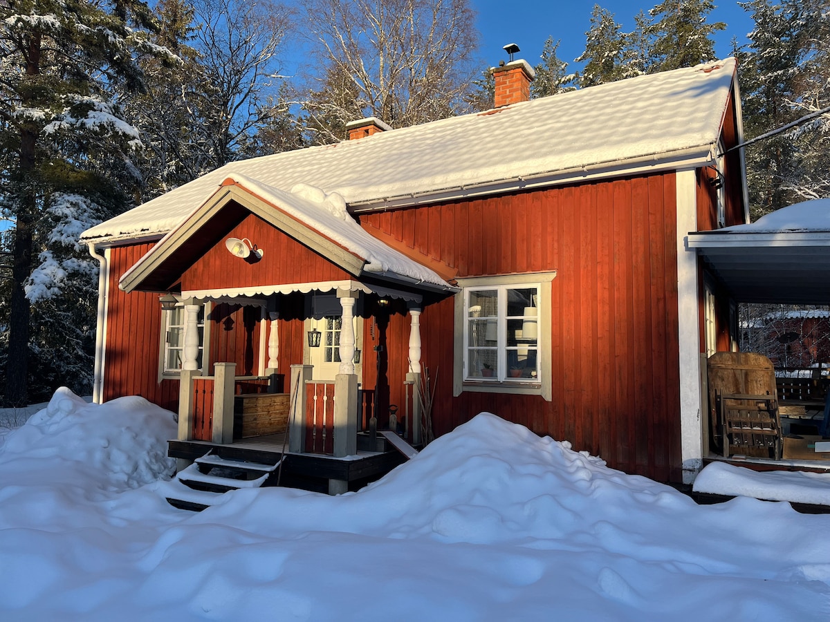 The exterior of a traditional red wooden house is depicted, surrounded by snow-covered ground. A small porch is highlighted with decorative elements. Snow blankets the landscape, and trees are visible in the background, contributing to the serene winter setting.