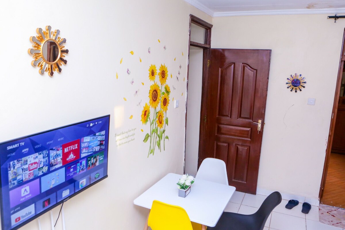 A compact dining area features a small table with two chairs, set against a wall adorned with sunflower decals. A wall-mounted television displays streaming options, with a decorative mirror reflecting natural light, and slippers are positioned near the entrance.