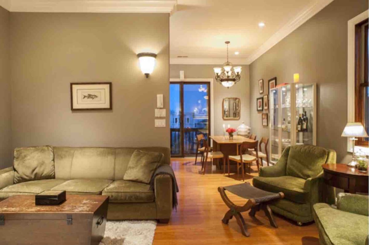 A tastefully arranged living area features a green sofa and a wooden coffee table. A dining table surrounded by chairs is visible in the background, with a side cabinet displaying glassware. Warm light illuminates the space from various fixtures and a nearby window.