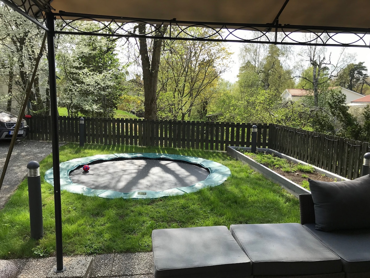 A grassy outdoor area is surrounded by a wooden fence, featuring a trampoline partially shaded by a canopy. Lush trees and blooming flowers provide a natural backdrop, while a lounging area with grey seating creates a comfortable space to relax.