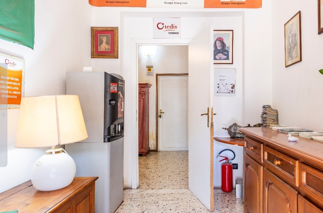 A well-lit entryway features a wooden cabinet and a light-colored lamp beside a black refrigerator. Artwork hangs on the walls, providing visual interest. A door leads to an interior space, and essential safety equipment is visible, contributing to a secure environment.