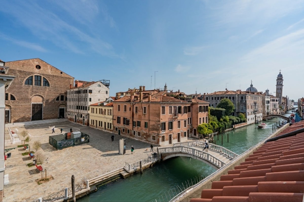 A panoramic view showcases a serene canal lined with classic Venetian buildings. A quaint bridge crosses the water, while historical structures with terracotta roofs and charming facades add character to the landscape. The sky above is bright and clear, enhancing the scene.