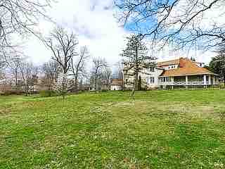 A spacious outdoor area is shown, featuring a lush green lawn dotted with mature trees. The exterior of a multi-storied house is visible in the background, surrounded by nature, suggesting a serene and inviting environment.