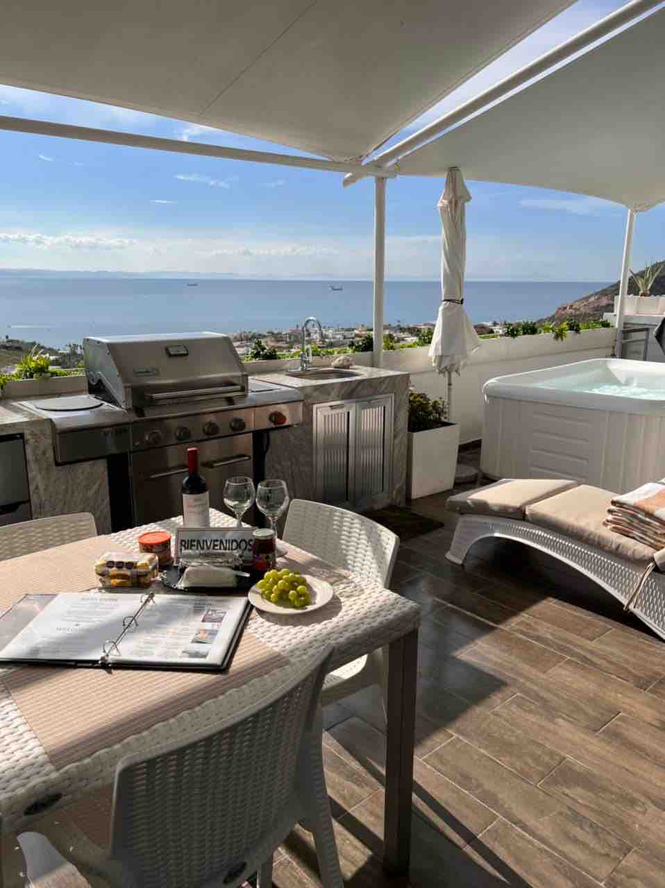 An outdoor terrace features a grill station, a hot tub, and a dining table set for four. Sun loungers are positioned nearby, with a view of the ocean in the background. The table displays a bottle of wine, glasses, and a plate of grapes.
