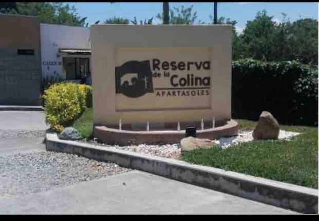 The entrance sign for Reserva de la Colina is displayed prominently. It features a neutral color palette and is surrounded by well-maintained landscaping, including green shrubs and decorative rocks, providing a welcoming first impression.