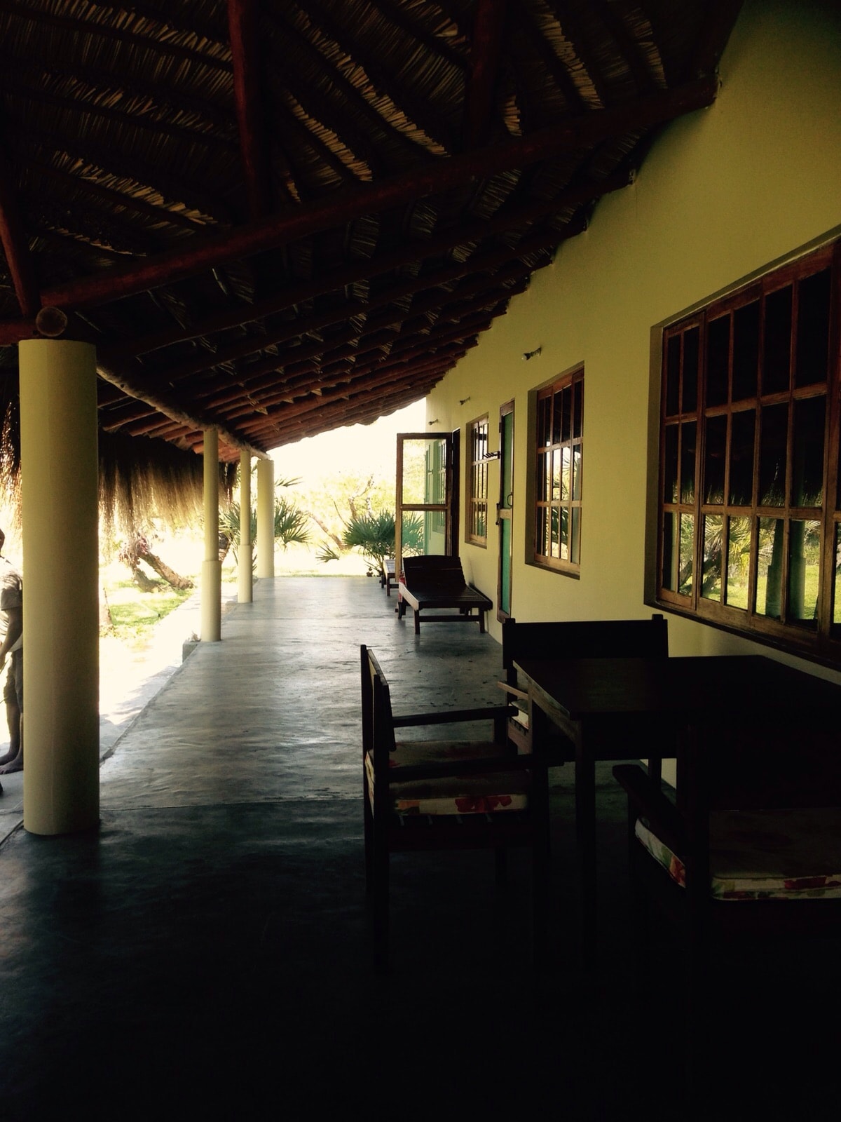A covered patio area is shown, featuring a long walkway with chairs and small tables. Large windows with wooden frames allow natural light to illuminate the space. A door leads outside to a garden setting, creating a sense of openness and connection to nature.