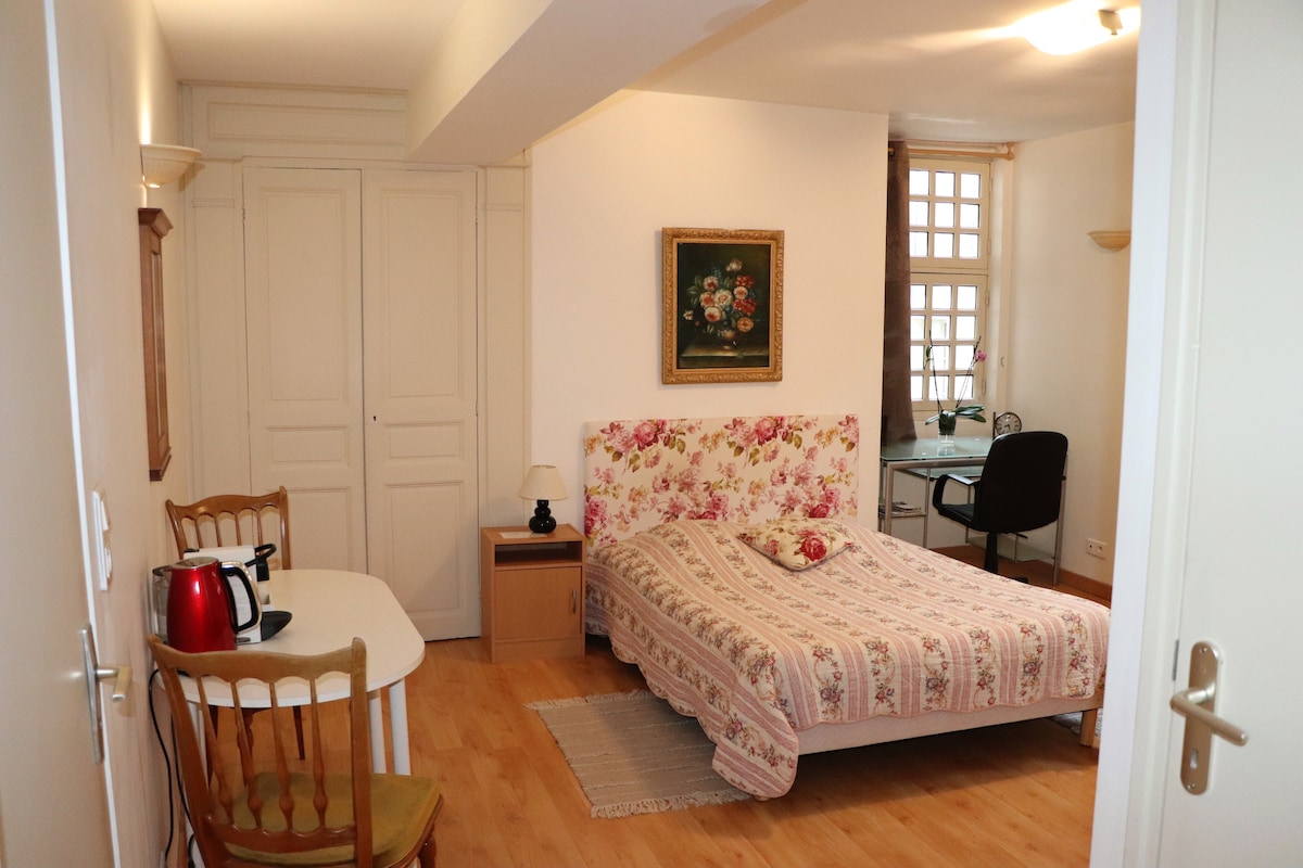 A private room features a floral-patterned bedspread on a spacious bed, complemented by a wooden table and chair set. Natural light enters through a window, while an art piece adorns the wall. A work desk with a chair is positioned in the corner.
