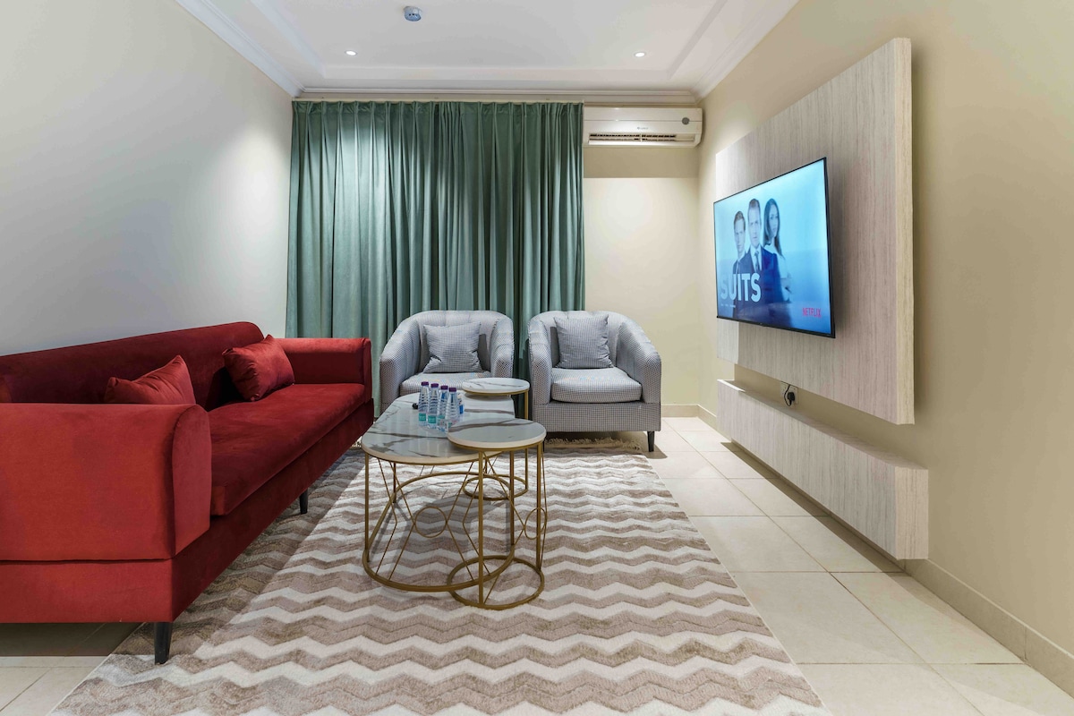 The living room features a red sofa and two gray armchairs, arranged around a round coffee table. A television is mounted on the wall opposite the seating area. Natural light is softened by green curtains, enhancing the room's comfortable ambiance.