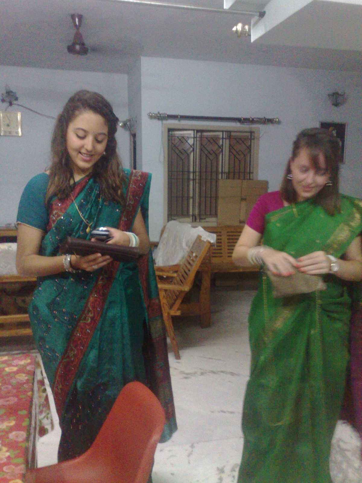 Two individuals are present in a room adorned with light-colored walls and a marble floor. One person is wearing a blue and green sari, while the other is dressed in a green sari. A wooden chair is visible in the background, alongside a table with a tablecloth.