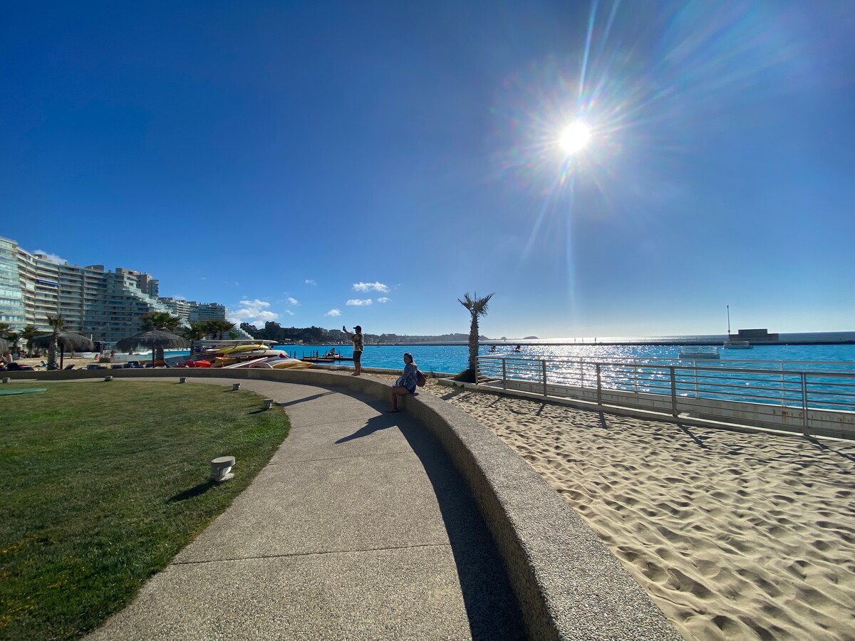 A scenic pathway runs alongside the beach, bordered by vibrant green grass and sandy areas. Calm waters reflect the sunlight, while kayaks and boats are visible in the distance. Palm trees provide shade, and a person is seated on the curved concrete wall enjoying the view.