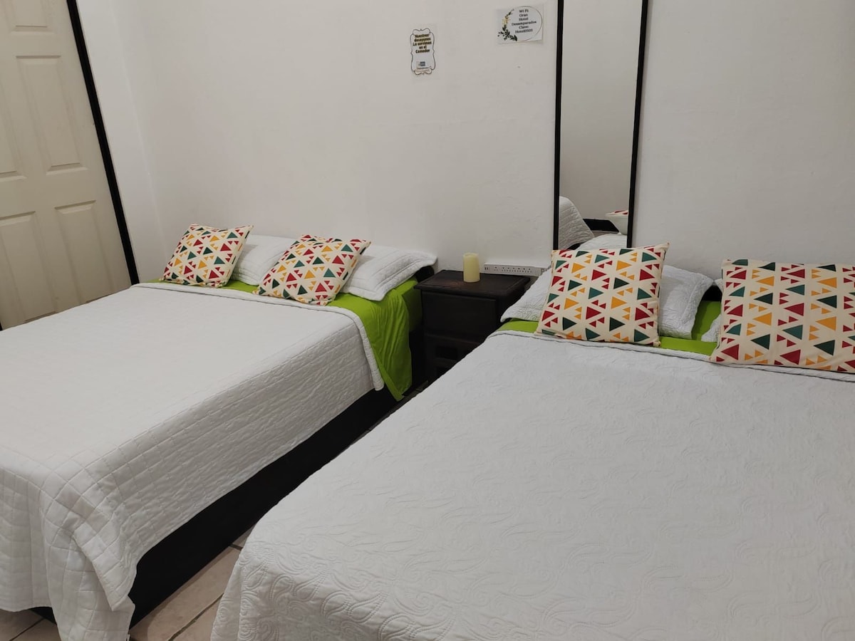 The family room features two spacious beds, each adorned with decorative pillows. A vibrant green accent is visible on the bedspreads, and mirrored wardrobes complement the natural light. A small nightstand with a candle is positioned between the beds, enhancing the functional layout.