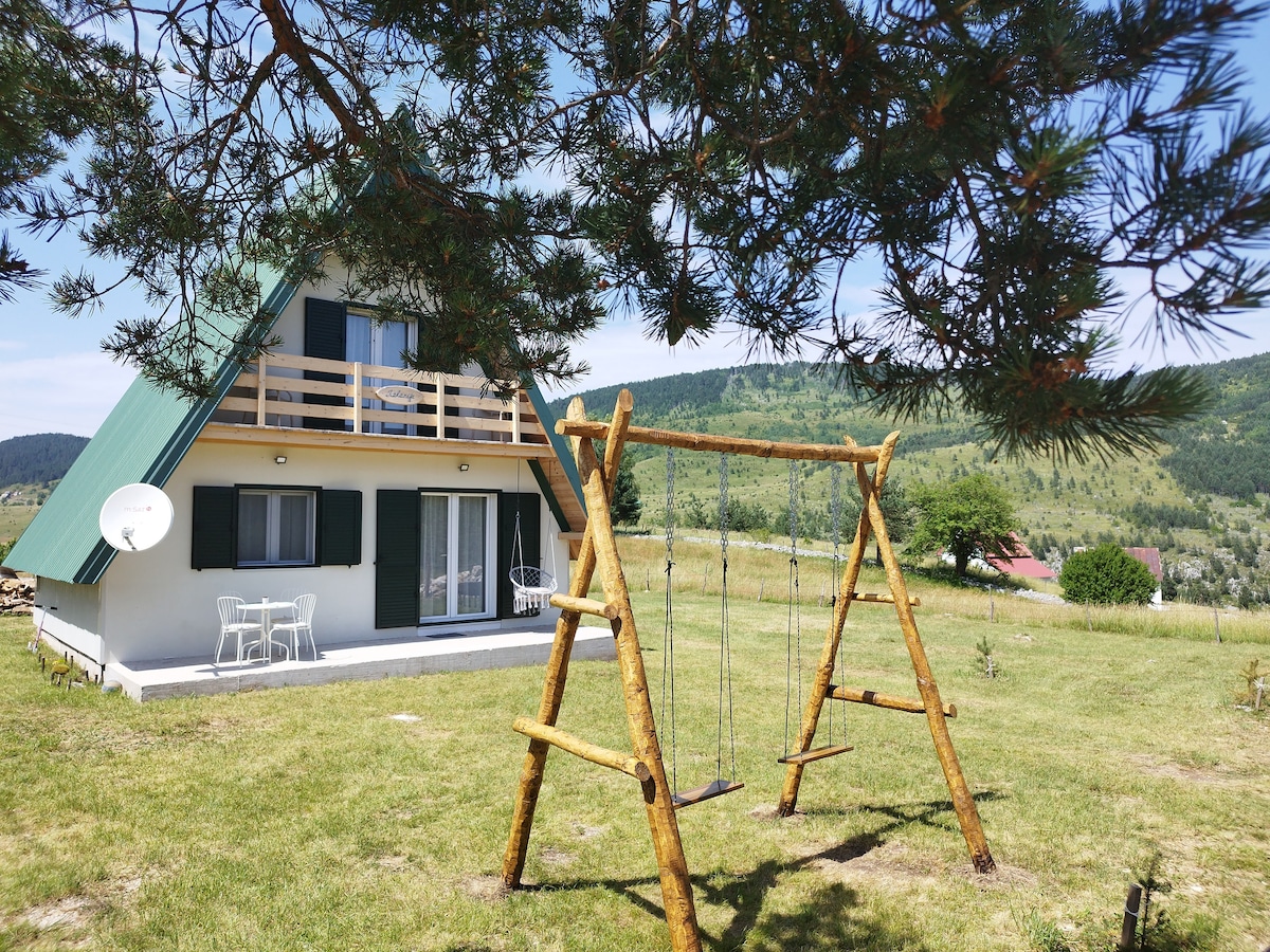 A wooden swing set is positioned on a grassy area in front of a two-story house. The house features a peaked green roof, white walls, and a terrace with outdoor seating. Lush green hills surround the property, providing a serene natural backdrop.