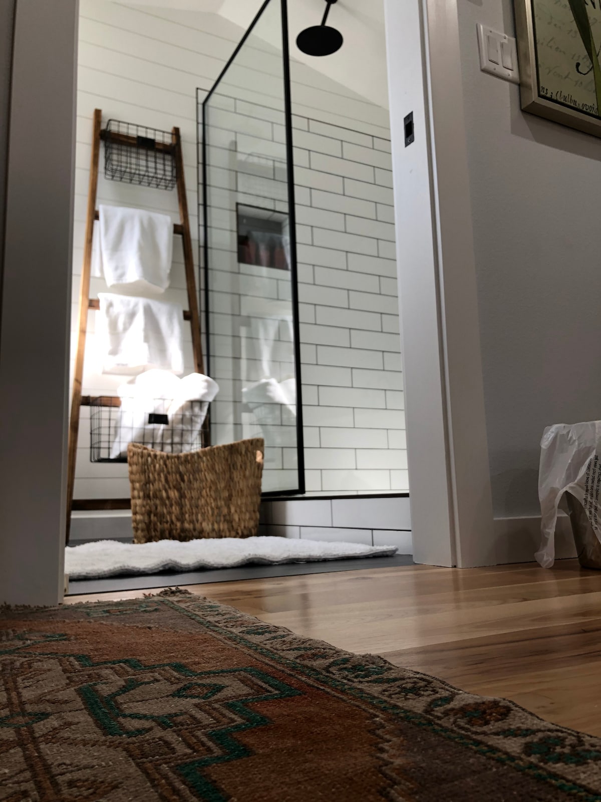 A modern shower room is featured, showcasing a glass-enclosed shower and white subway tile walls. A wooden ladder displays fluffy white towels, while a woven basket sits on a soft rug outside the shower. Natural light brightens the space.