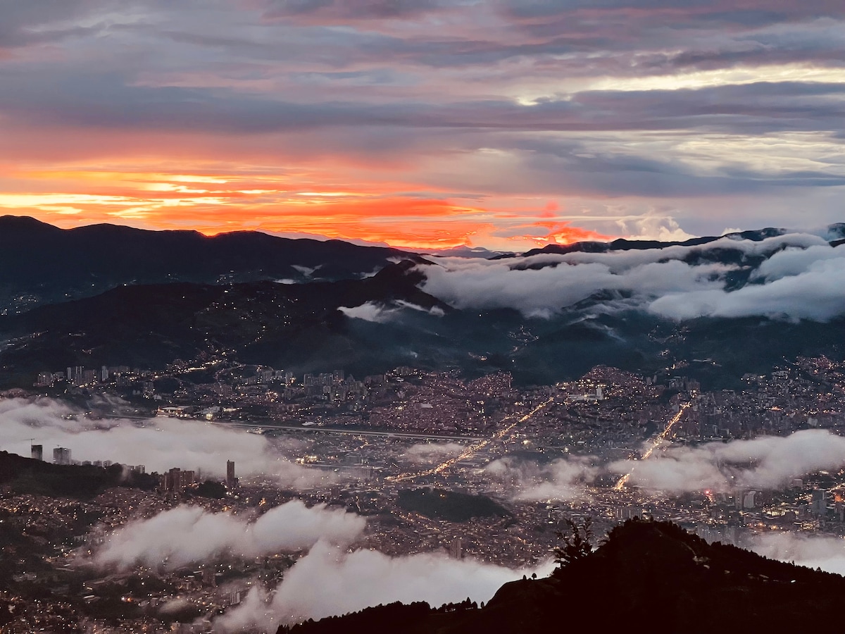 A panoramic view of Medellin at sunset, showcasing the city's twinkling lights and the surrounding mountains. Clouds blanket the lower areas, creating a serene atmosphere as the sky transitions from orange to soft blue hues.