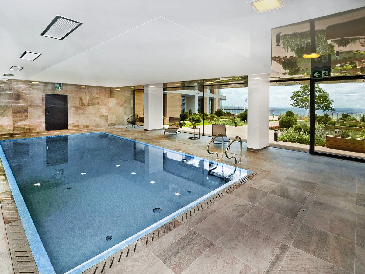 An indoor pool area is visible, featuring a gleaming blue pool surrounded by sleek, smooth tiles. Comfortable seating is positioned alongside the water, with large windows that provide views of the outdoor landscape. Natural light enhances the open and modern feel of the space.