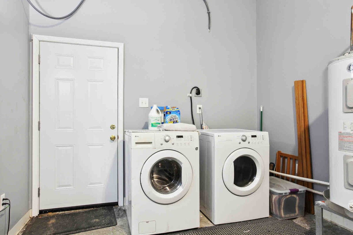 A laundry area is equipped with a pair of front-loading washing machines positioned beside a door leading outside. The space features gray walls and a textured gray floor. Supplies are organized on a shelf above the machines, contributing to a functional and streamlined environment.
