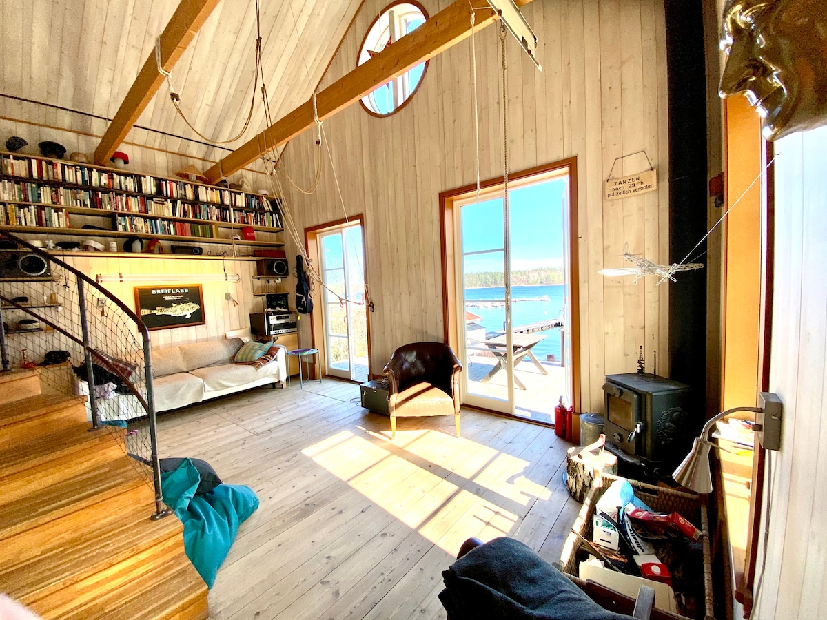 A spacious living area features wooden walls and high ceilings with exposed beams. Large windows allow natural light to fill the room, while a plush couch and cozy chair provide seating options. A wood-burning stove and bookshelves enhance the inviting environment.