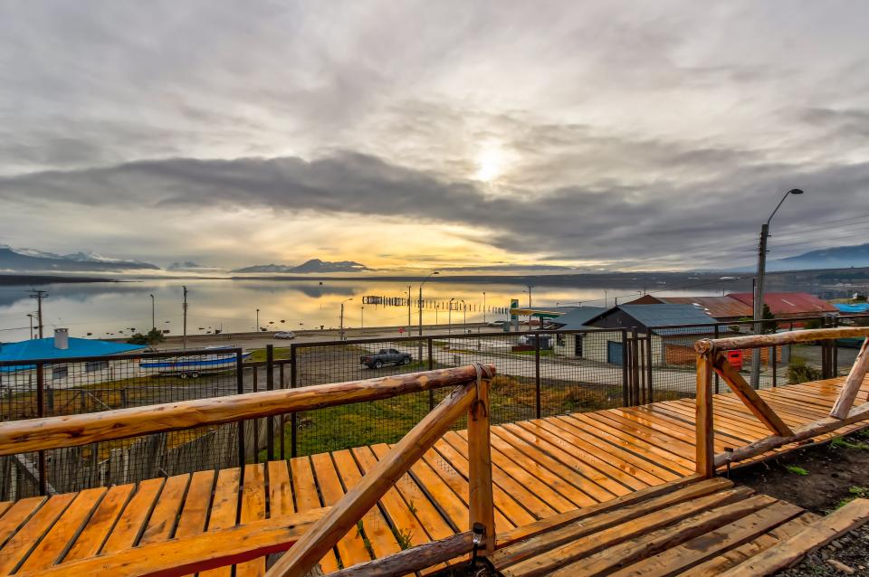 A wooden pathway leads toward a serene waterfront view, where calm waters reflect the soft colors of an overcast sky. Industrial structures and a distant pier are visible along the shoreline, with the silhouette of mountains in the background.