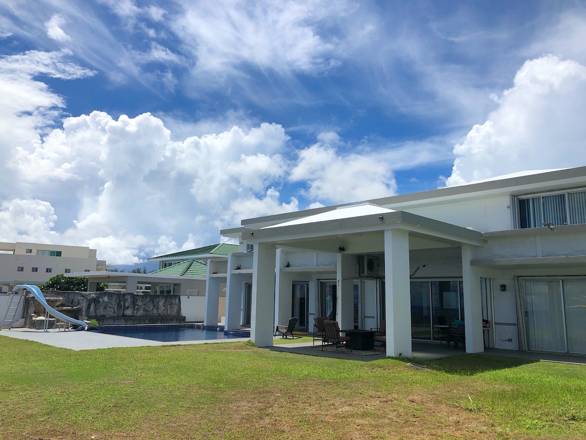 The exterior of a two-story home is shown, featuring a spacious backyard with a swimming pool and a water slide. Lounge chairs are positioned on the patio, and the sky is filled with fluffy clouds above. Green grass surrounds the pool area.