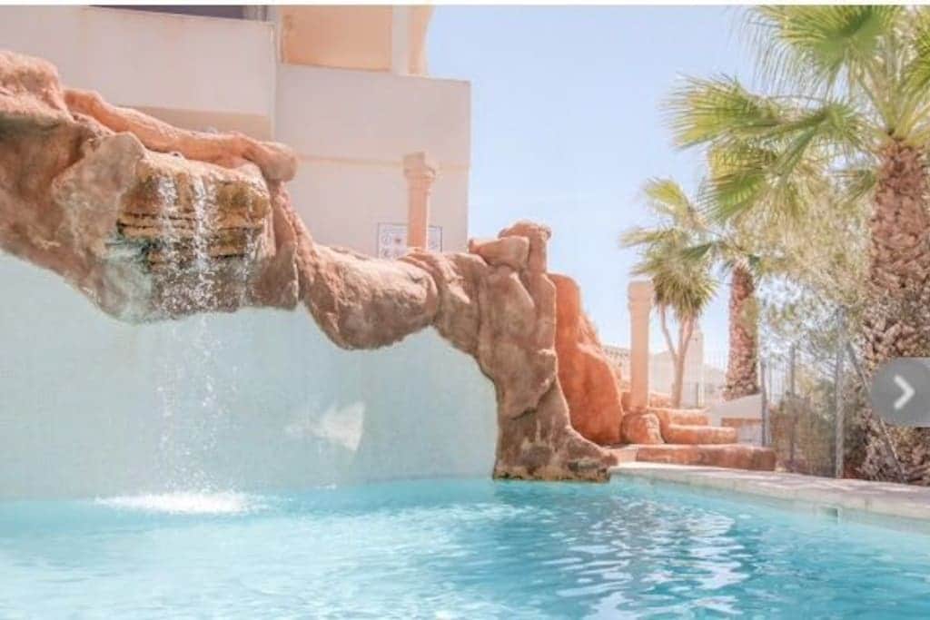 A tranquil pool is framed by a rock formation, featuring a gentle waterfall. Lush palm trees are visible in the background, enhancing the serene atmosphere. Sunlight reflects off the water's surface, creating a bright and inviting space for relaxation.
