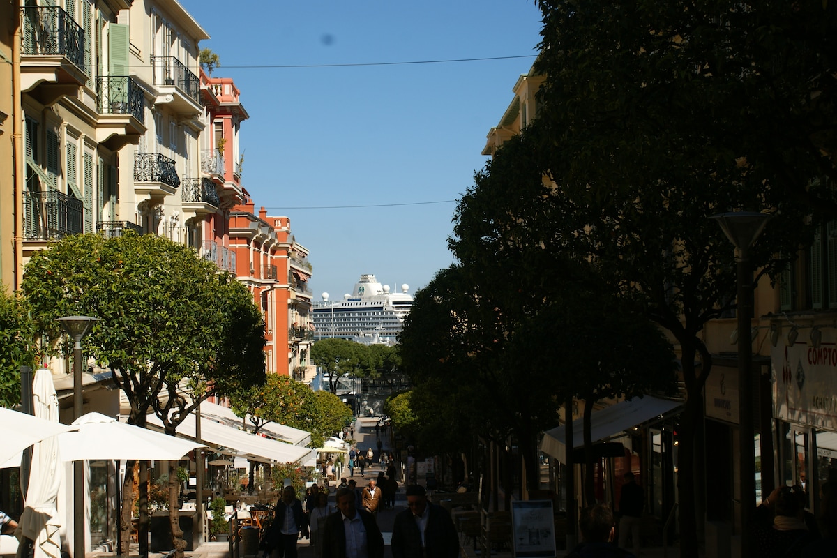 A vibrant street scene is captured, lined with trees and colorful buildings. Cafe tables are arranged along the walkway, inviting passersby. In the distance, a cruise ship can be seen docked, adding a lively element to the bustling atmosphere.