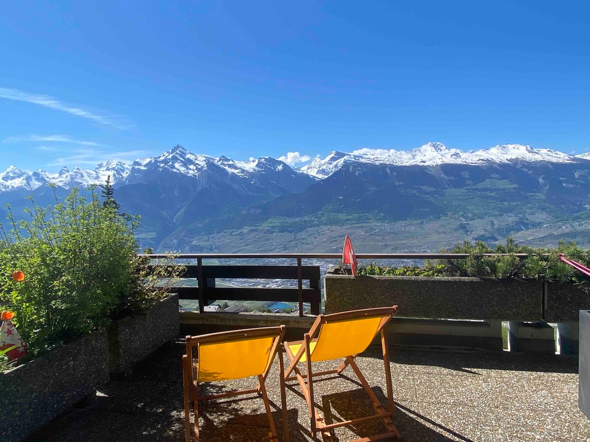 A balcony is shown with two wooden lounge chairs facing a breathtaking mountain view. Snow-capped peaks rise in the distance, framed by a clear blue sky. Green plants are seen along the balcony's edge, adding a touch of nature to the serene setting.