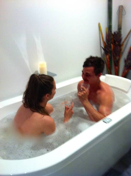 A spacious bathtub is filled with bubbles, creating a relaxing atmosphere. A candle is positioned nearby, adding a soft glow. Decorative elements include bamboo accents in the background, contributing to a serene setting for unwinding.