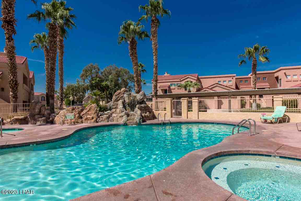 A sparkling pool is surrounded by palm trees and a rocky feature, providing a refreshing retreat. Adjacent to the pool are lounge chairs, and a hot tub is visible in the background, creating a relaxing outdoor space within the complex.