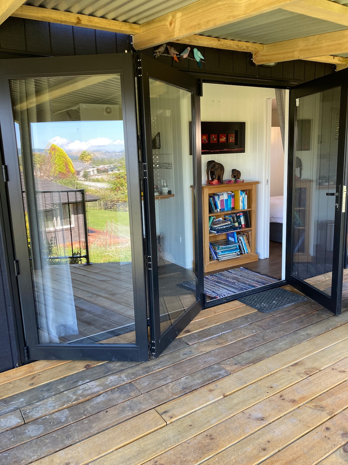 Double glass doors open from a wooden deck into a cozy interior space. A small bookshelf filled with various books is visible, alongside subtle decorative elements. In the background, a glimpse of a bedroom can be seen, with soft natural light illuminating the area.