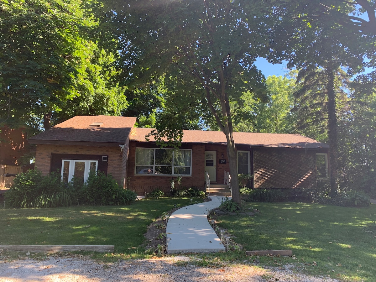 A single-story bungalow is surrounded by lush greenery, featuring a well-maintained path leading to its entrance. Large windows allow natural light to illuminate the inviting façade, while varied landscaping adds to the exterior's charm.