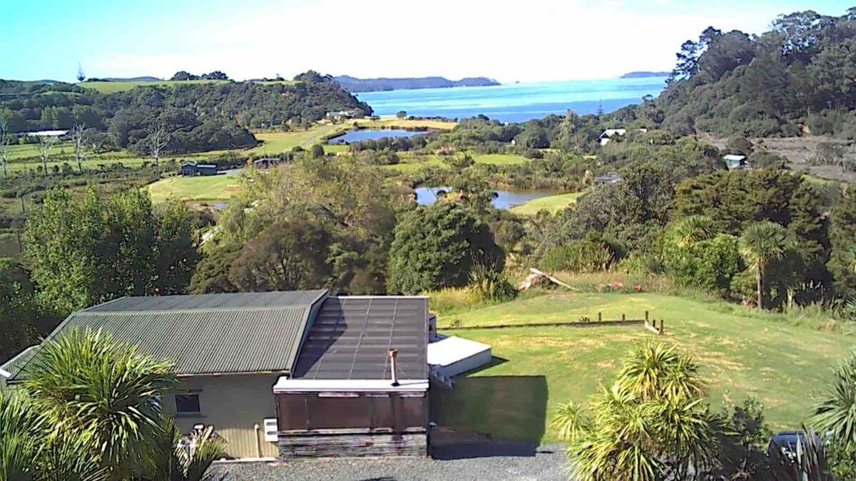A peaceful view captures the surrounding landscape, featuring lush greenery, rolling hills, and shimmering water in the distance. The cottage's roof is visible on the left, while palm trees and other foliage frame the scene, creating a sense of tranquility and connection to nature.
