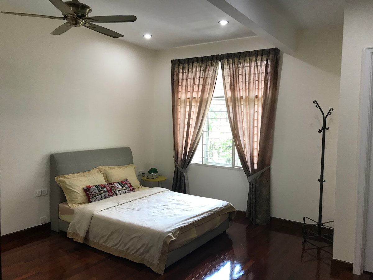 A well-lit bedroom features a spacious bed dressed in neutral colors. Two large windows allow natural light to fill the room, complemented by beautiful drapes. A small side table and a coat rack are present, adding practical elements to the inviting space.