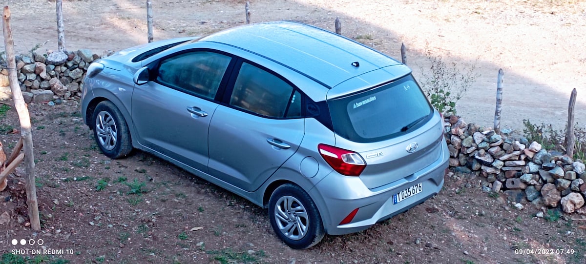 A silver compact car is parked on a gravel surface, partially on a slight incline. Surrounding the vehicle is a natural landscape, with a stone boundary visible in the background.