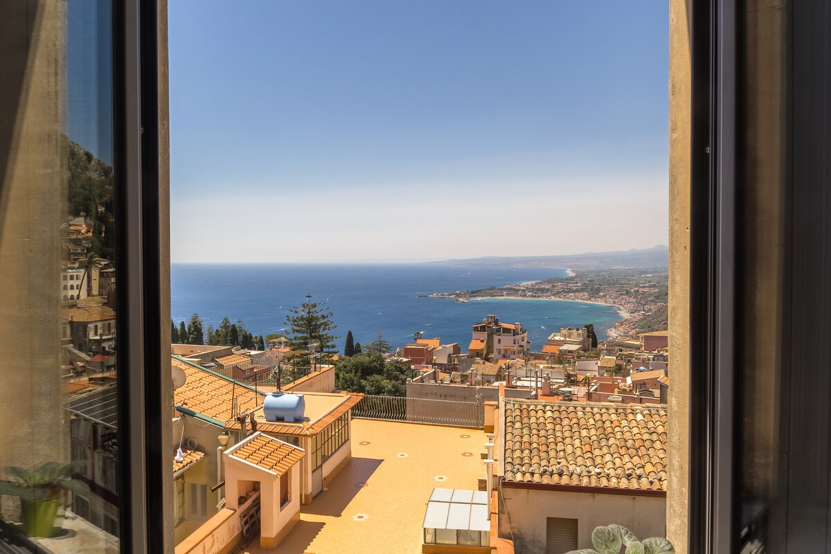 A panoramic view is captured through large windows, revealing the coastline and the blue sea beyond. The surrounding architecture includes terracotta roofs and greenery, with clear skies adding to the serene ambiance. The horizon stretches into the distance, showcasing a tranquil seaside landscape.
