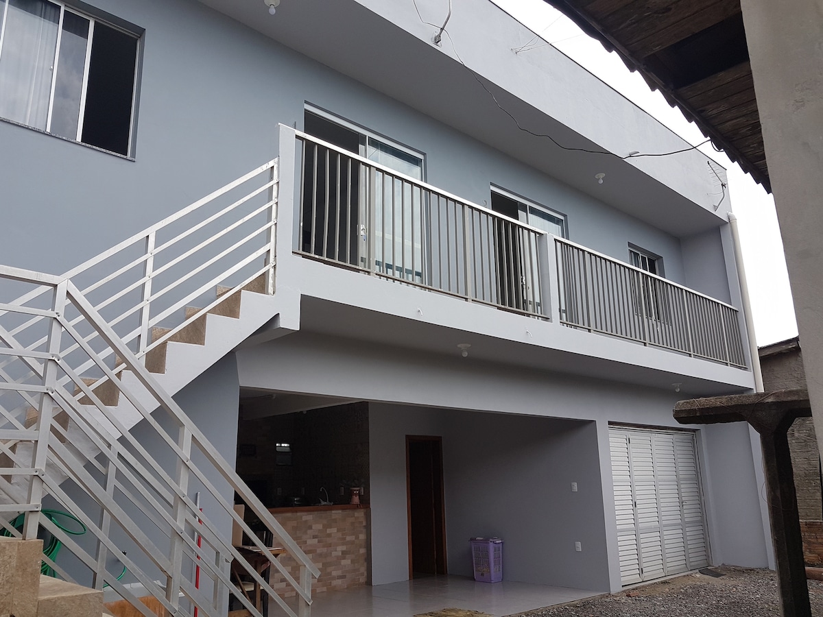 The exterior of a multi-level building is highlighted, featuring a light gray facade and a wide balcony with railings. An outdoor staircase leads to the upper level, while the open area below includes a small kitchen visible through glass doors.