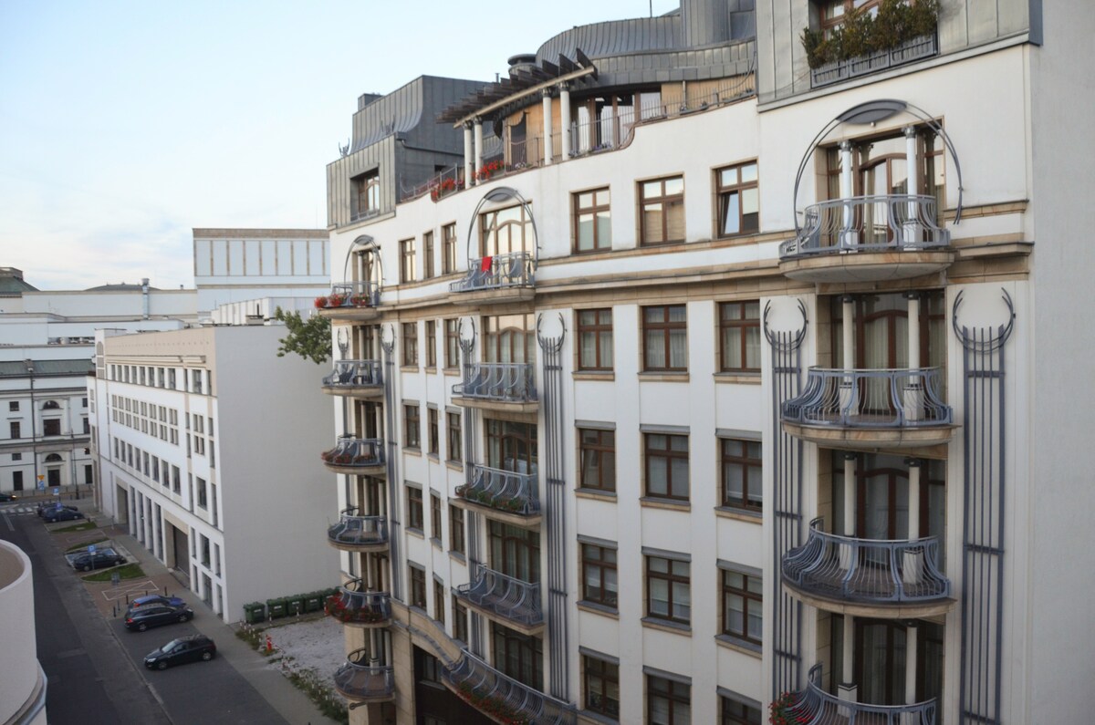 A multi-story building showcases a blend of modern architectural elements and classic design. Balconies extend from several windows, adorned with decorative railings. The surrounding area features additional buildings, and the sky reflects soft hues of evening light.
