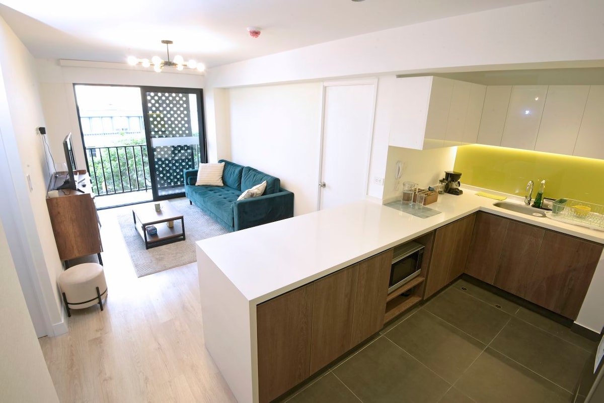 A modern living area showcases a comfortable green sofa facing a wall-mounted television. Natural light enters through a balcony door, leading to a small outdoor space. The kitchenette features sleek finishes, equipped with essential appliances, including a countertop and cabinets.