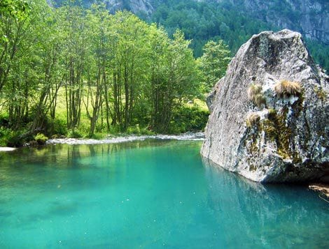 A serene natural scene showcases a calm body of water reflecting vibrant turquoise hues, bordered by lush greenery and trees. A large boulder with moss sits prominently at the water's edge, adding texture to the tranquil setting.
