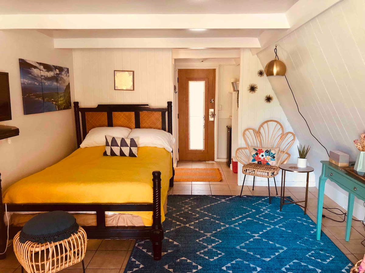 A bright sleeping area is highlighted by a spacious bed with a yellow cover. A cozy seating area features a woven chair and a decorative side table. The room's decor includes artwork and a potted plant, with natural light entering through a wooden door.