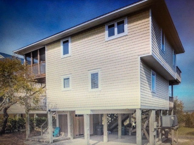 The exterior of the house is elevated on stilts, featuring a pale yellow wooden facade. Windows are positioned symmetrically, allowing for natural light. An outdoor area beneath the structure is visible, providing shaded space with some seating and storage options.