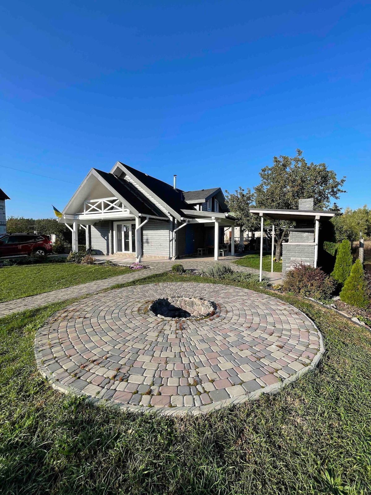 The exterior of a two-story wooden house is presented, featuring a stone-paved circular area with a fire pit at its center. Lush greenery surrounds the home, and a covered structure is visible, providing shaded space. Clear blue skies complete the serene outdoor setting.